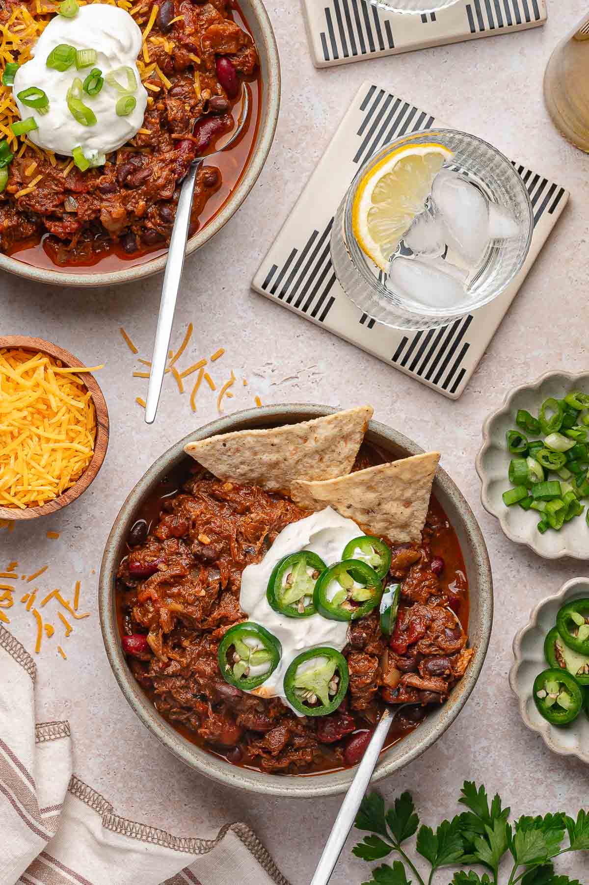 Two bowls of pulled pork chili topped with sour cream, shredded cheese, sliced jalapeรฑos, green onions, and tortilla chips, served with drinks and garnishes on a light table.