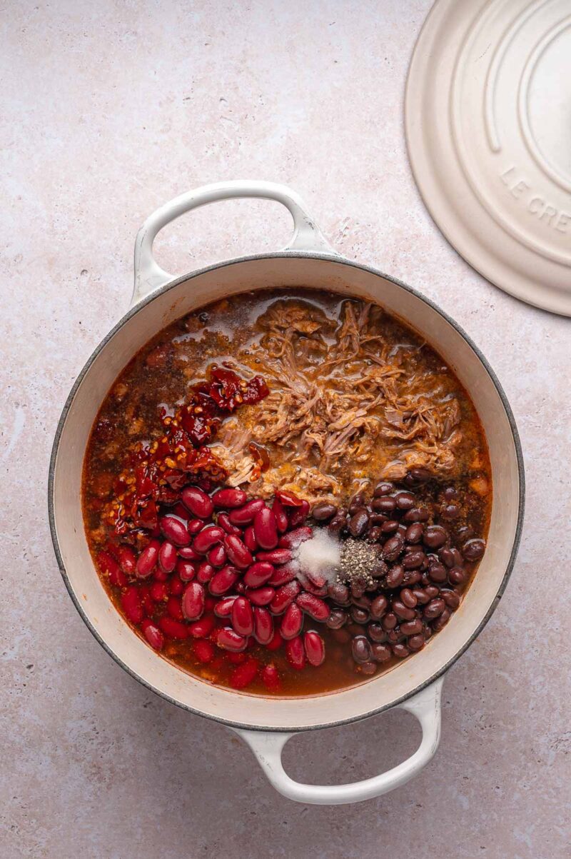 A white pot filled with pulled pork chili—shredded meat, kidney beans, black beans, spices, and broth—ready to be mixed for cooking; the lid rests beside the pot.