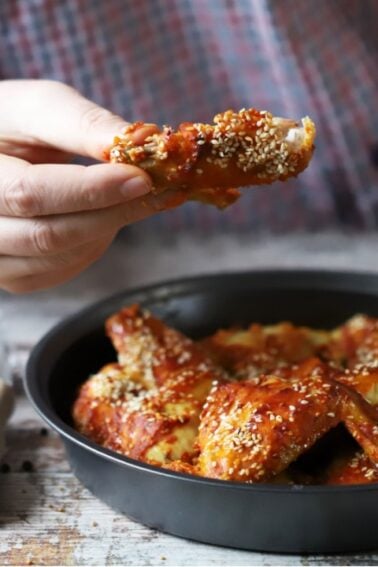 Person holding a saucy, sesame-coated chicken wing above a black dish filled with similar wings; condiments and spices are arranged on the table nearby.