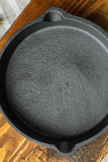 A small, empty cast iron skillet on a wooden cutting board, with a striped cloth, bay leaf, and rosemary sprig nearby.