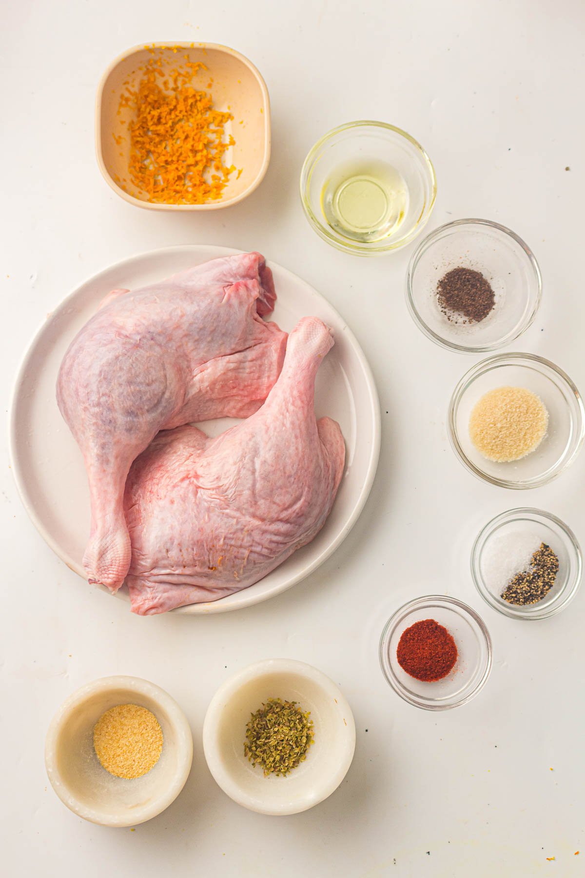 Raw duck legs on a plate surrounded by small bowls containing oil, grated orange zest, salt, pepper, paprika, garlic powder, onion powder, and dried herbs on a white surface.