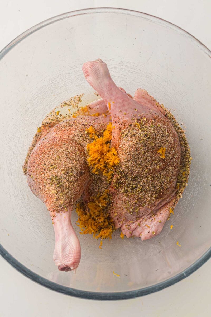 Two raw duck legs in a glass bowl coated with a spice mixture and orange zest.