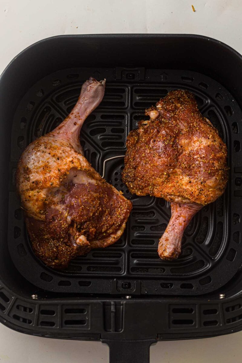 Two seasoned duck legs placed side by side in the basket of an air fryer, ready for cooking.