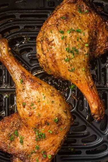 Two seasoned, air fryer duck legs with crispy skin rest on an air fryer basket, garnished with chopped herbs.