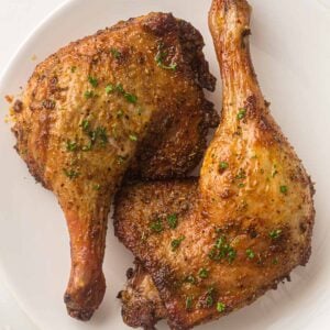 Two cooked and seasoned duck leg quarters with crispy skin served on a white plate, garnished with chopped herbs.