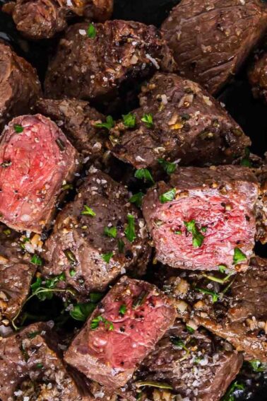 A black plate filled with cooked steak bites, some pieces showing a pink center, garnished with chopped herbs and coarse salt.