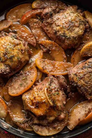Skillet with cooked chicken thighs, sliced apples, and herbs in a brown sauce on a wooden table, with salt, pepper, and a whole apple nearby.
