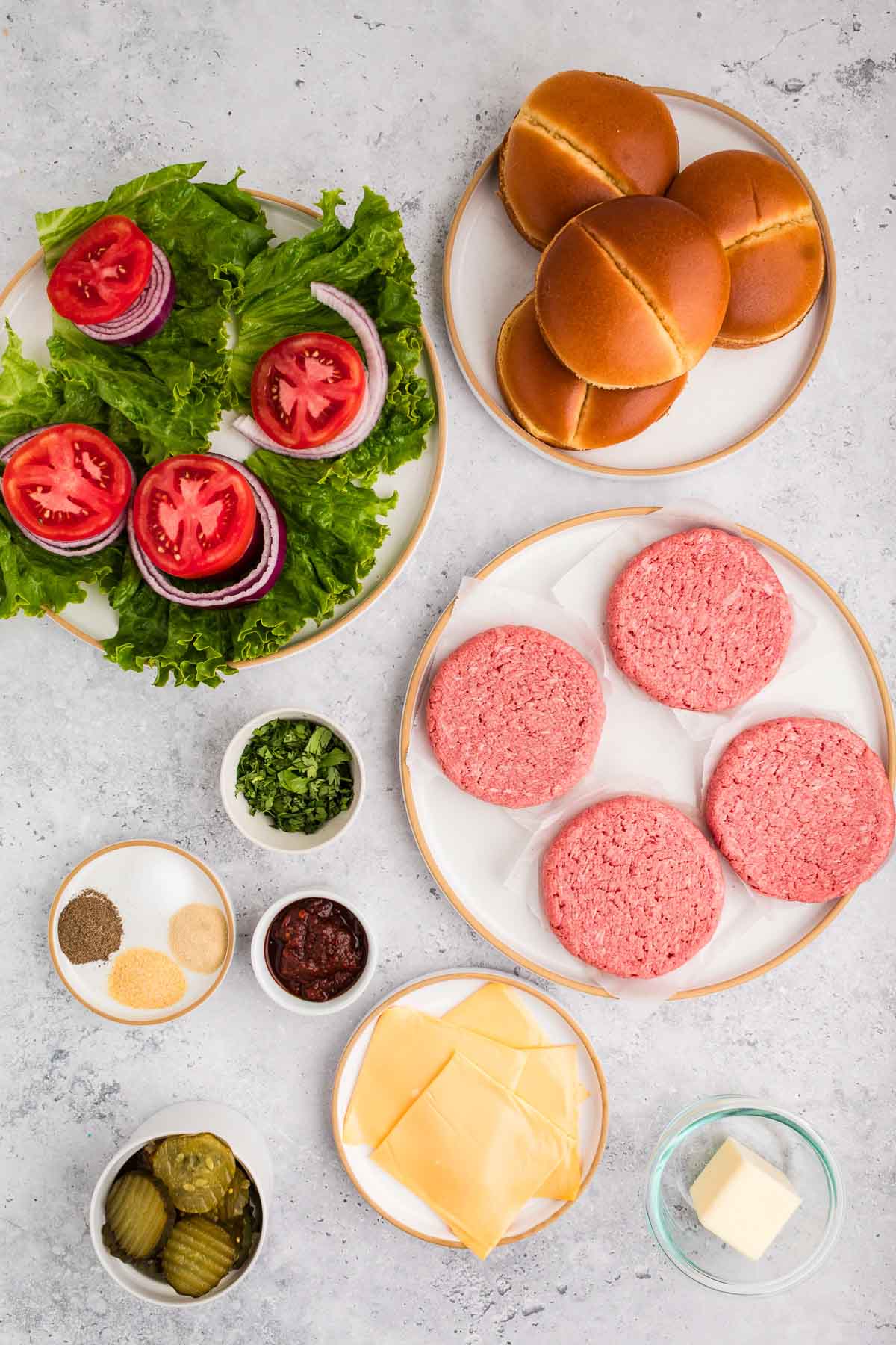Burger ingredients arranged on a table, including raw beef patties, buns, lettuce, tomato, onion, sliced cheese, pickles, seasonings, chopped herbs, sauce, and a small bowl of butter.