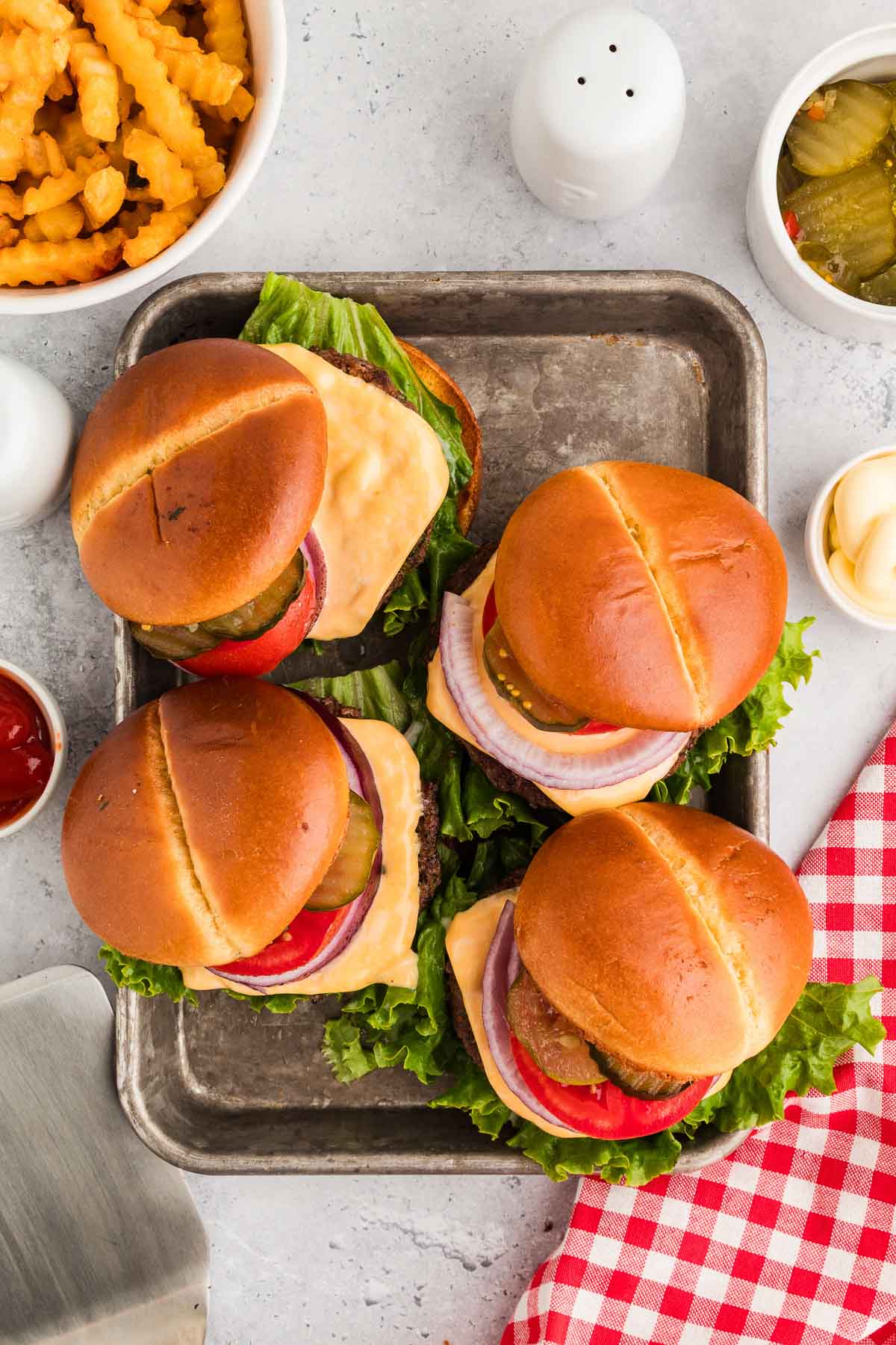 Four cowboy butter burgers with lettuce, tomato, pickles, onion, and cheese on a tray, surrounded by crinkle-cut fries, pickles, ketchup, mayonnaise, and a red checkered napkin.