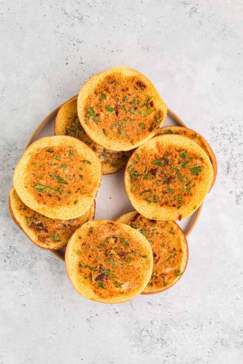 Four toasted sandwich buns with cowboy butter are arranged in a circle on a round plate, placed on a light gray textured surface.