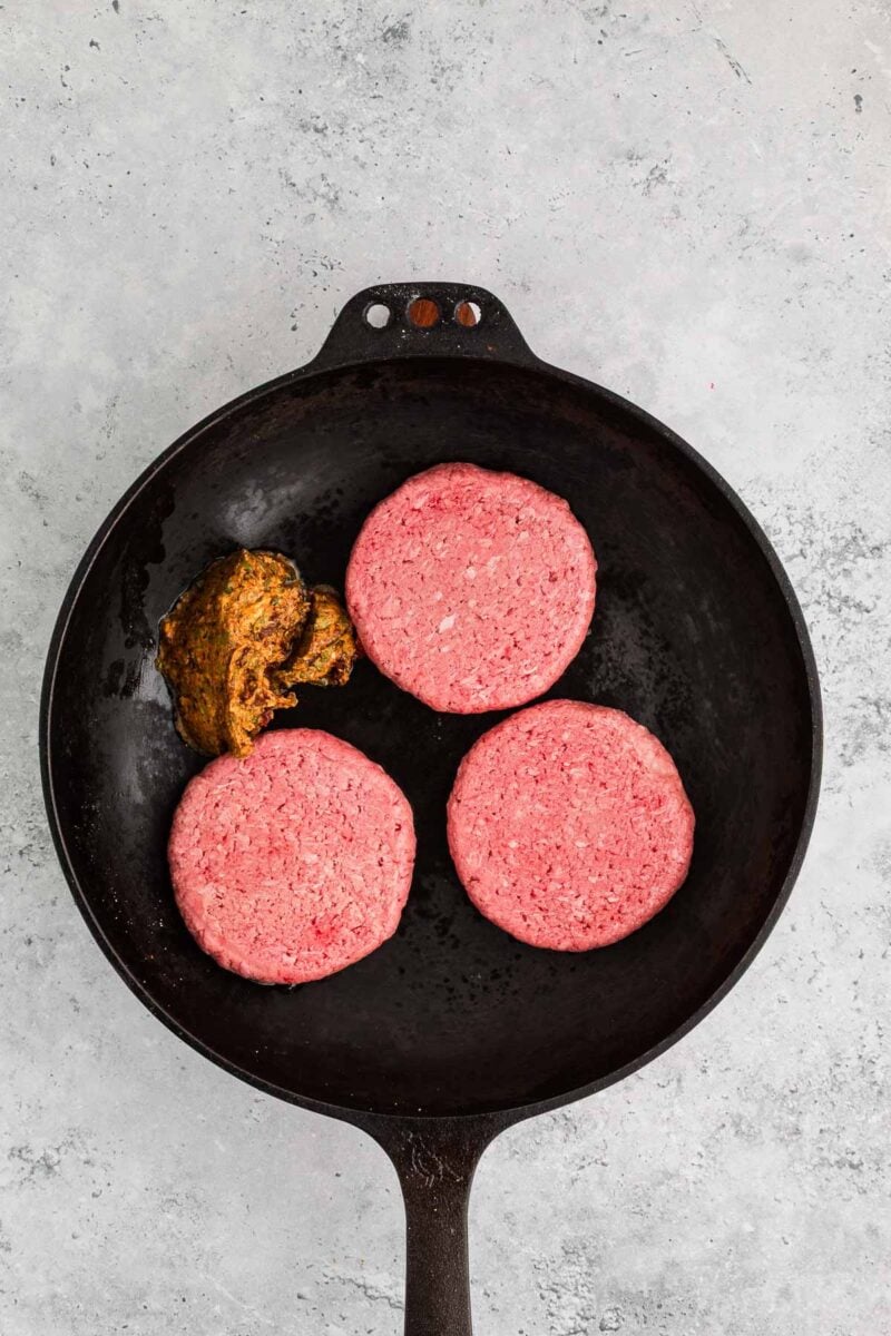 Three raw burger patties and a portion of cowboy butter are placed in a black skillet on a gray surface.