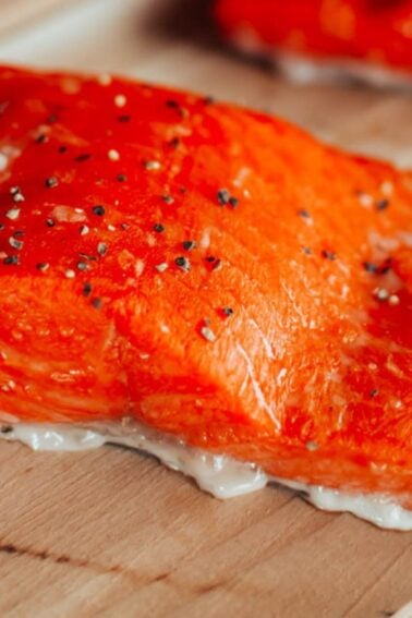 A raw salmon fillet with salt and pepper seasoning rests on a wooden plank.
