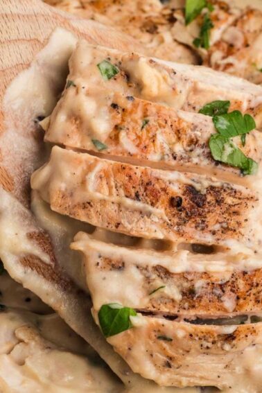Sliced cooked chicken breast in a creamy garlic sauce, garnished with chopped parsley, being lifted with a wooden spatula.