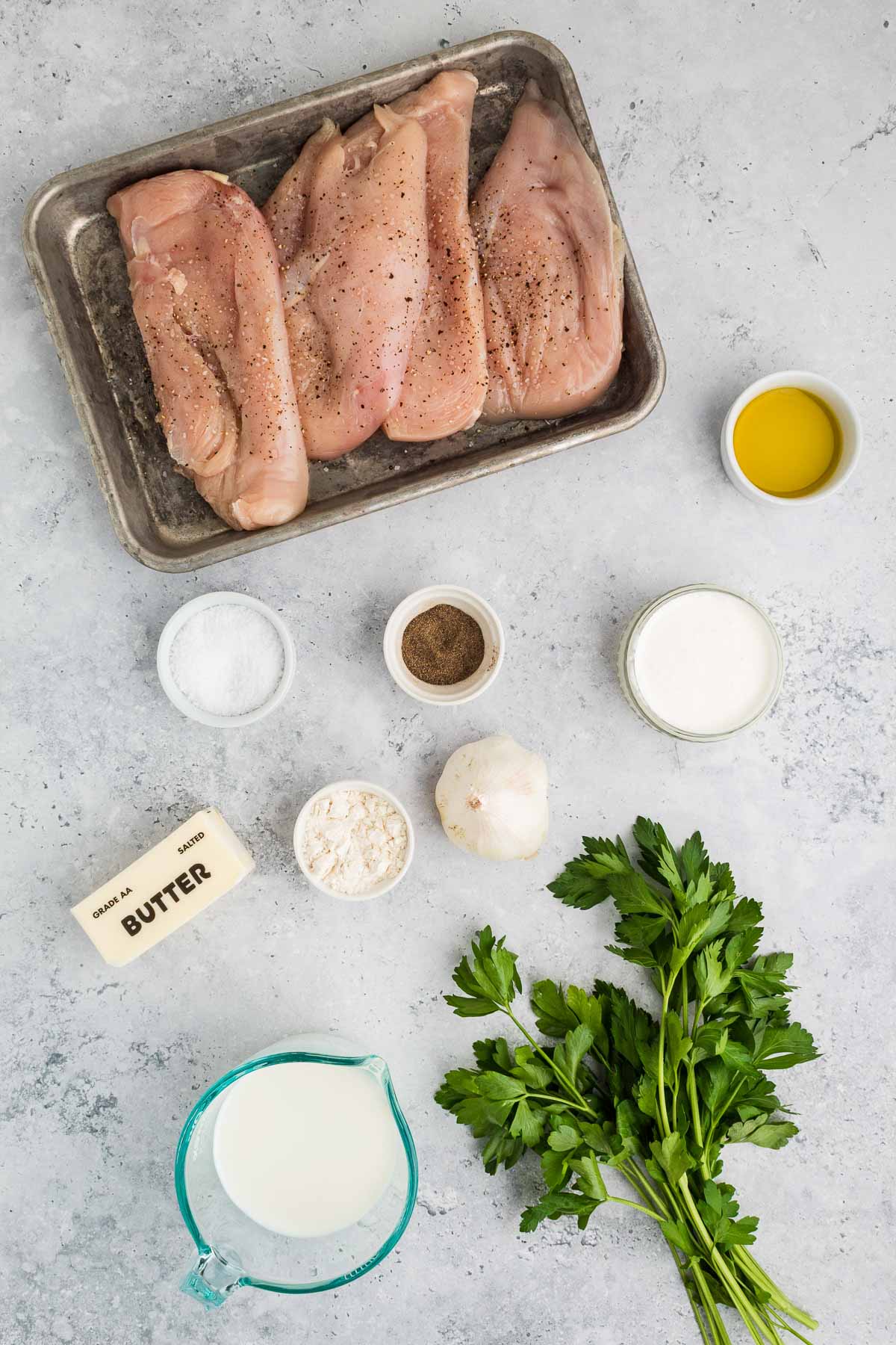 Raw seasoned chicken breasts on a tray, surrounded by small bowls of olive oil, salt, pepper, flour, milk, cream, butter, garlic, and fresh parsley on a gray surface.