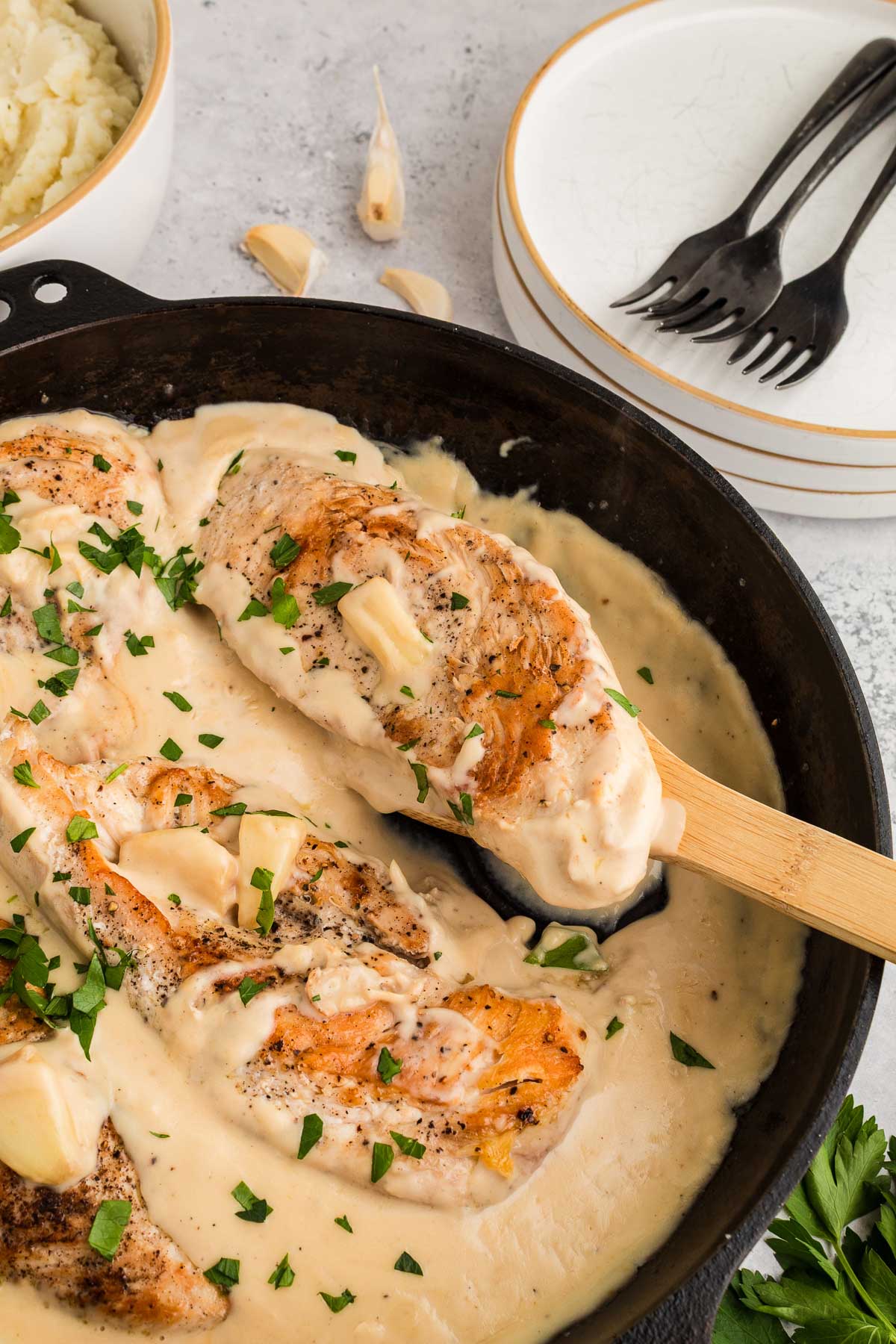 Seared chicken breasts in a creamy garlic sauce, garnished with chopped parsley, served in a black skillet with a wooden spatula. Plates, forks, and mashed potatoes in the background.