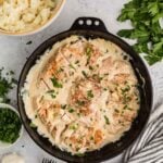 A cast iron skillet with creamy garlic chicken swimming in sauce, garnished with chopped herbs, beside mashed potatoes, fresh parsley, garlic, and salt and pepper shakers.