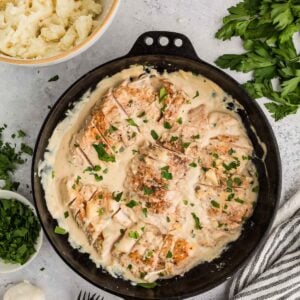 A cast iron skillet with creamy garlic chicken swimming in sauce, garnished with chopped herbs, beside mashed potatoes, fresh parsley, garlic, and salt and pepper shakers.