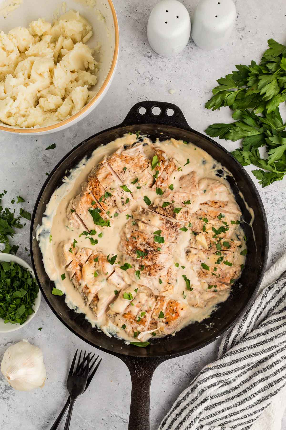 A cast iron skillet with creamy garlic chicken swimming in sauce, garnished with chopped herbs, beside mashed potatoes, fresh parsley, garlic, and salt and pepper shakers.