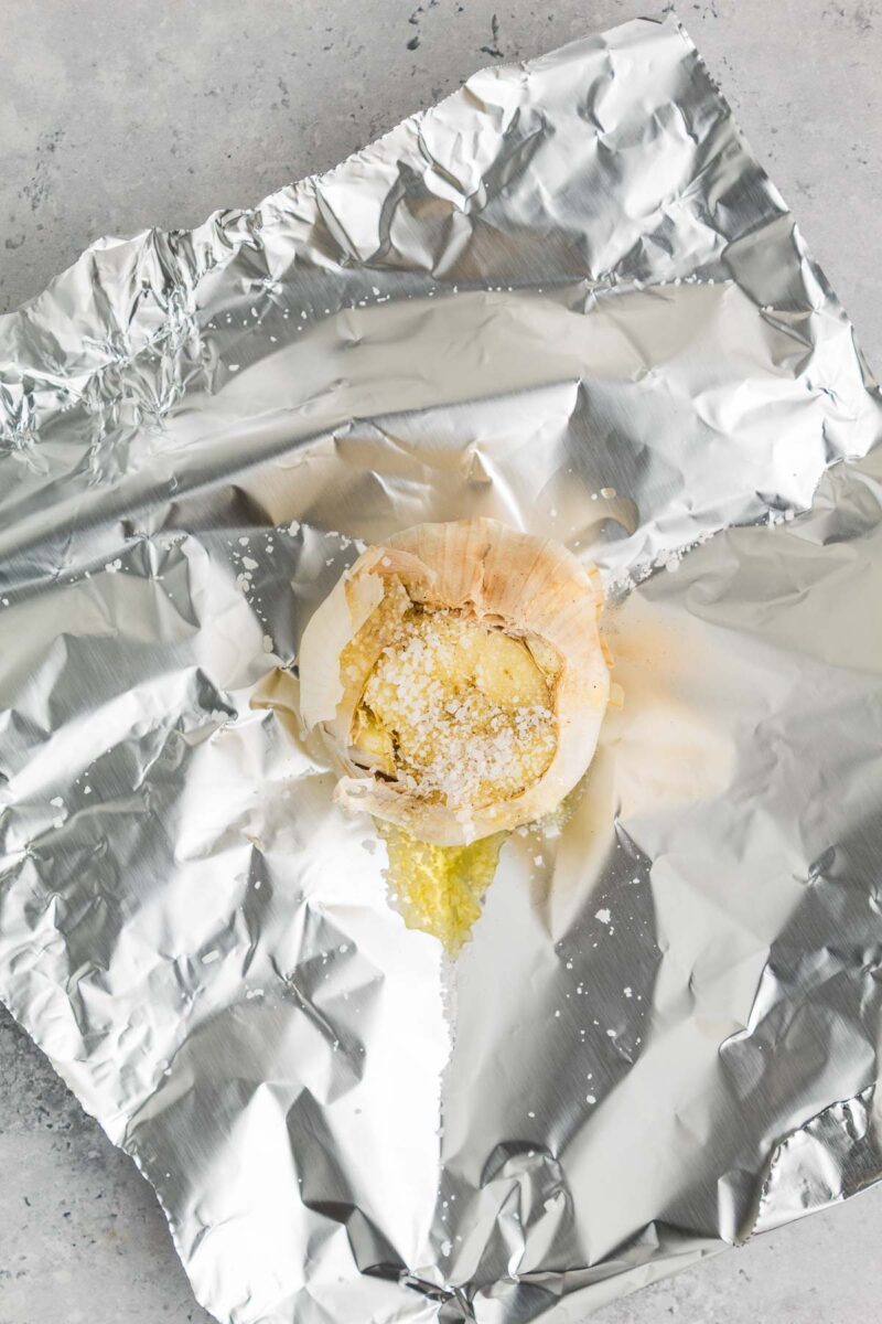 A head of garlic with oil and salt sits on a sheet of aluminum foil, ready to be wrapped for roasting.