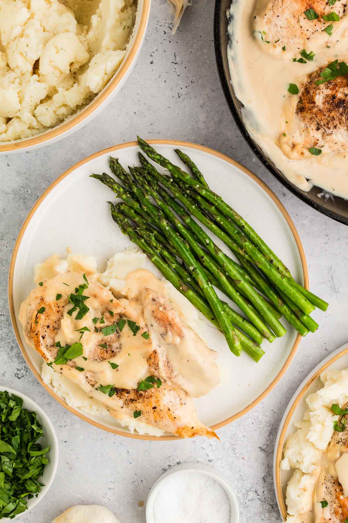 A plate with cooked chicken in creamy garlic sauce, roasted asparagus, and mashed potatoes, surrounded by bowls of mashed potatoes, chopped herbs, and salt.