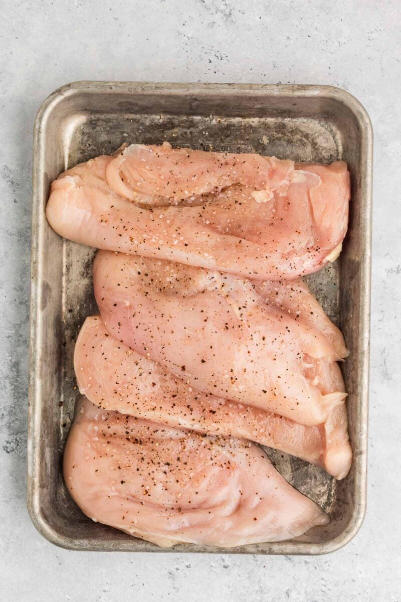 Four raw chicken breasts seasoned with salt and pepper are arranged on a metal baking tray against a light gray background.