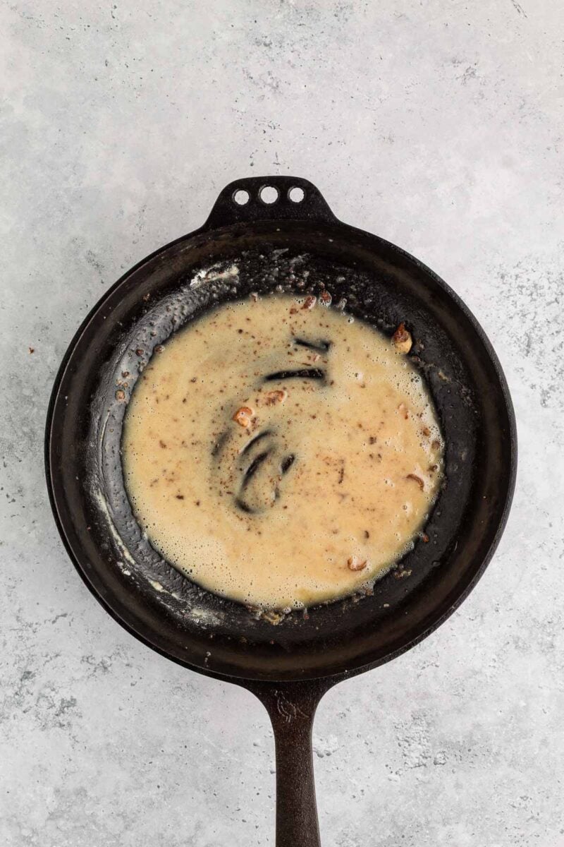 A cast iron skillet with a roux and browned bits on the bottom sits on a light gray surface.