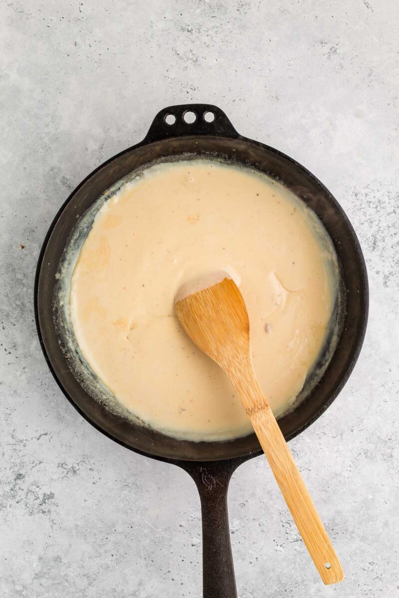 A cast iron skillet containing a chicken sauce sauce being stirred with a wooden spatula on a gray countertop.