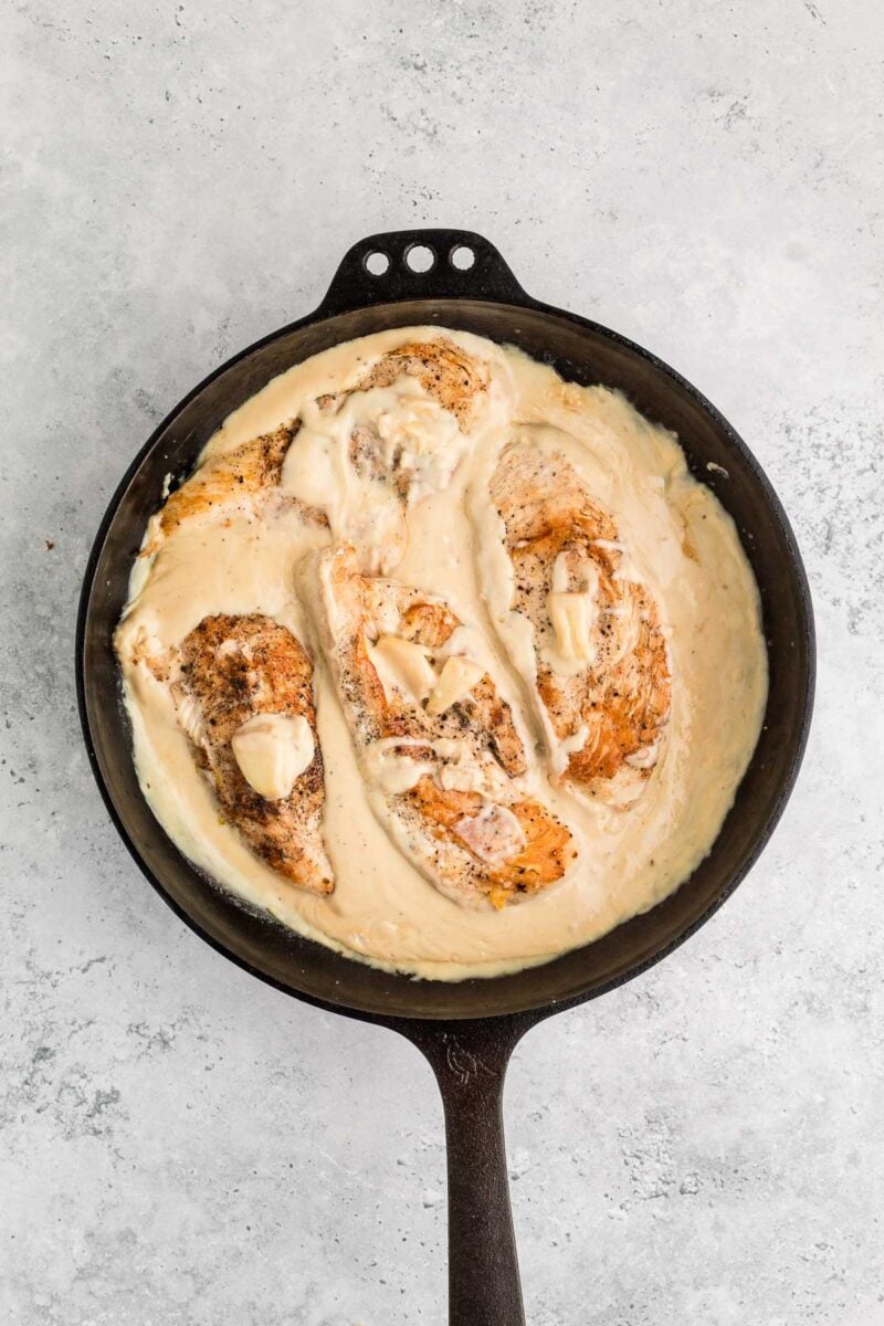 Cast iron skillet with browned chicken breasts in a creamy garlic sauce on a light gray surface.