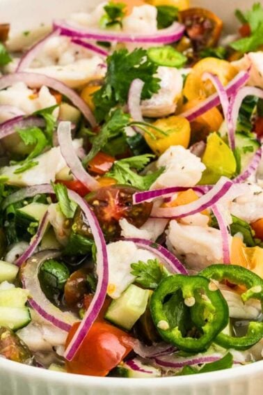 A bowl of ceviche with shrimp, diced tomatoes, cucumbers, red onion, cilantro, jalapeรฑo slices, and avocado slices on top.