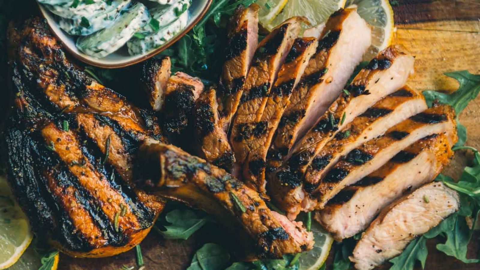 Grilled pork chops sliced and served on a wooden board with fresh greens, lemon slices, and a small bowl of herb sauce.