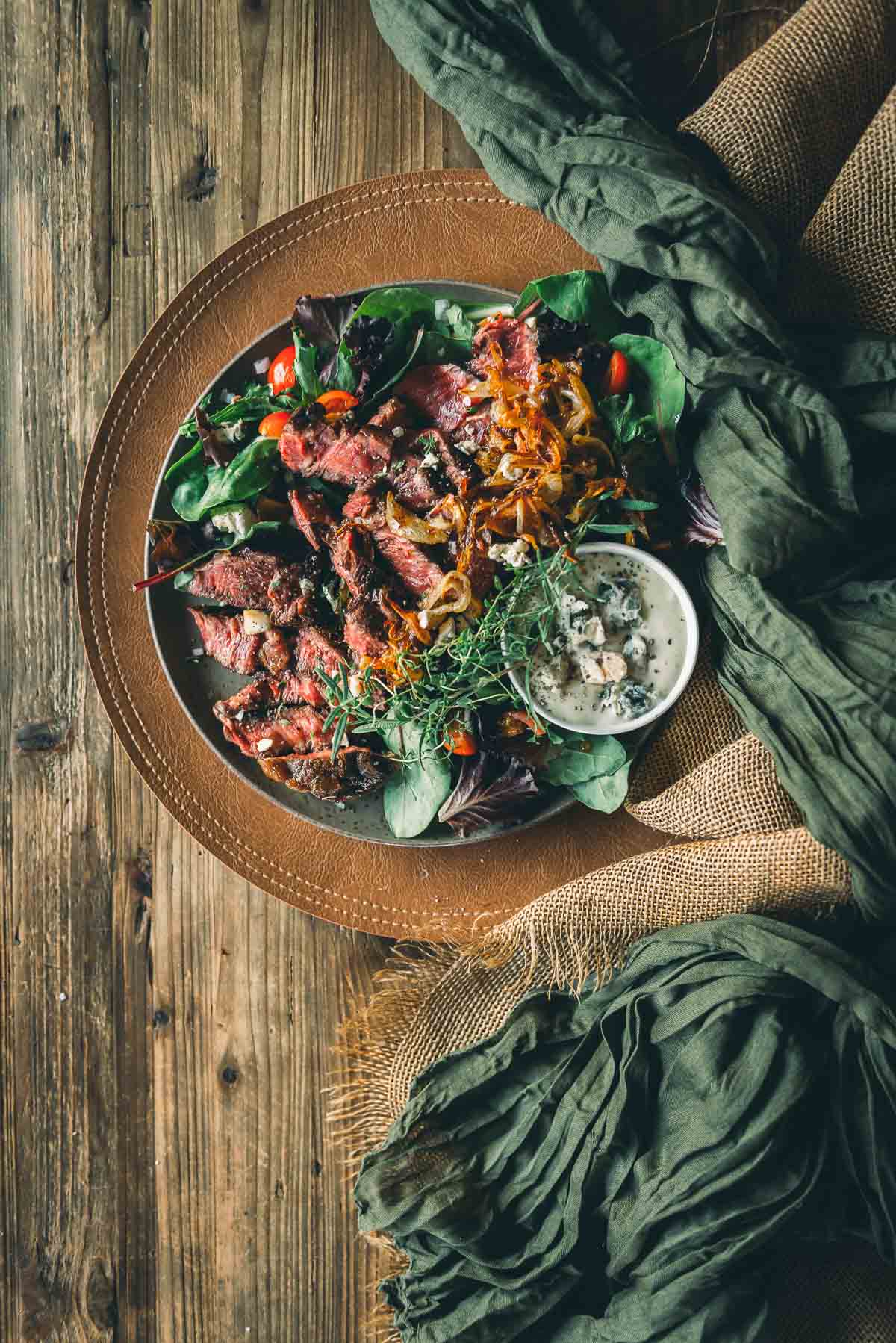 A plate of sliced chuck steak on mixed greens with roasted onions, cherry tomatoes, microgreens, and a small bowl of creamy dressing, set on a rustic wooden table.
