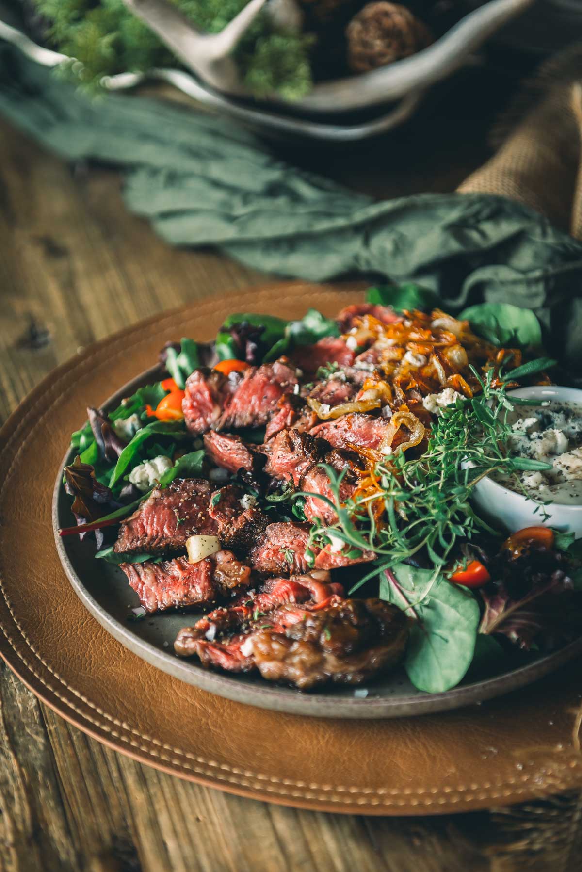 A plate of sliced seared chuck eye steak served on mixed greens with caramelized onions, fresh herbs, and a small dish of blue cheese dressing on a wooden table.
