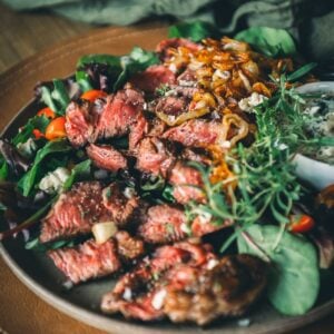 Plate of sliced beef chuck steak served on a bed of mixed greens, cherry tomatoes, caramelized onions, and crumbled cheese, garnished with fresh herbs.