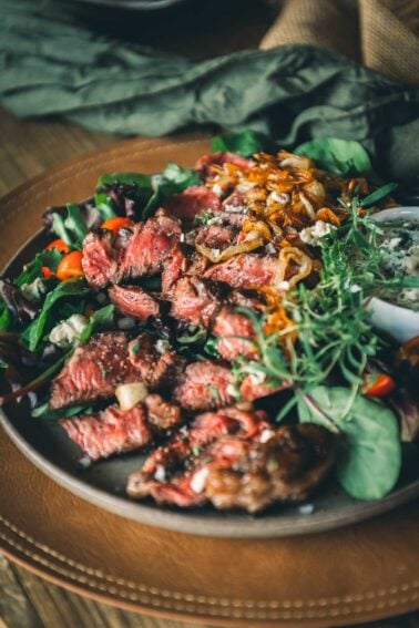 Plate of sliced beef chuck steak served on a bed of mixed greens, cherry tomatoes, caramelized onions, and crumbled cheese, garnished with fresh herbs.