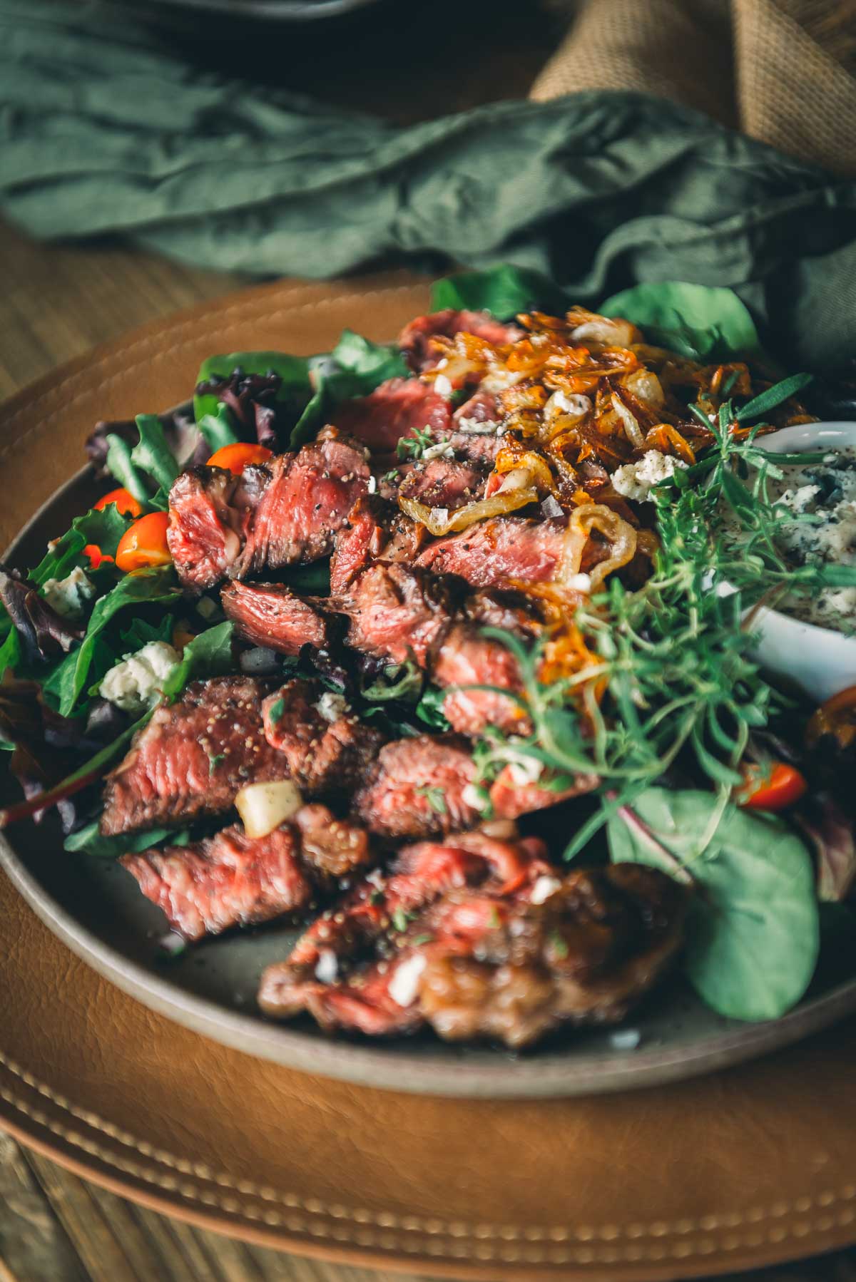 Plate of sliced beef chuck steak served on a bed of mixed greens, cherry tomatoes, caramelized onions, and crumbled cheese, garnished with fresh herbs.