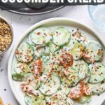 A bowl of cucumber salad with cream cheese, salmon, and seasoning, surrounded by chopsticks and small bowls of spices and sauce. Text overlay reads: “How to Make Philly Roll Cucumber Salad.”.