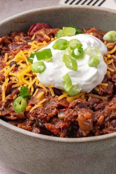 A bowl of pulled pork chili topped with shredded cheese, sour cream, and sliced green onions, with a bowl of shredded cheese and sliced jalapeños in the background.