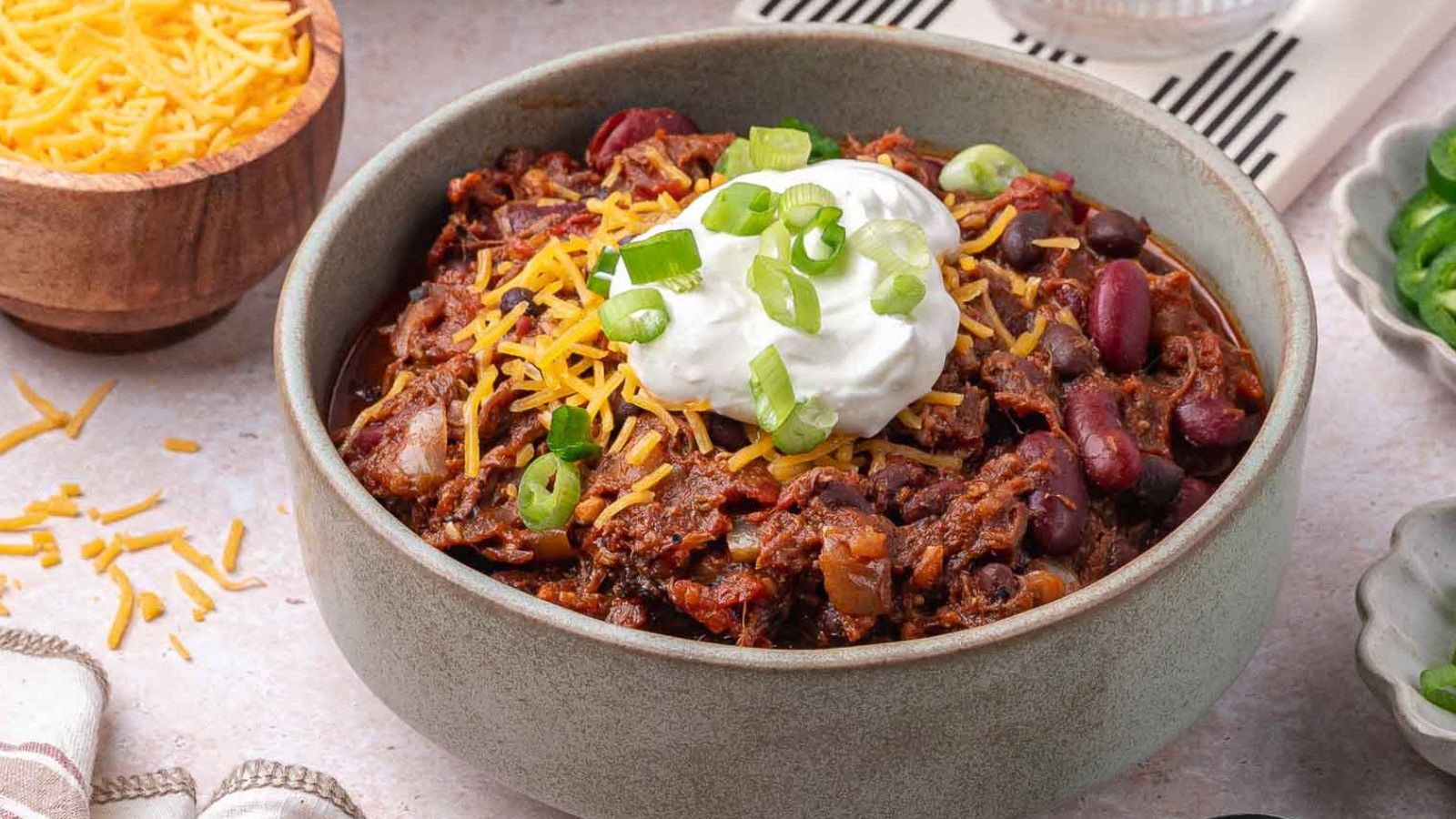 A bowl of pulled pork chili topped with shredded cheese, sour cream, and sliced green onions, with a bowl of shredded cheese and sliced jalapeños in the background.