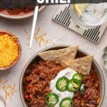 A bowl of pulled pork chili topped with sour cream, sliced jalapeños, and tortilla chips, with a side of shredded cheese and a spoon on a napkin nearby.