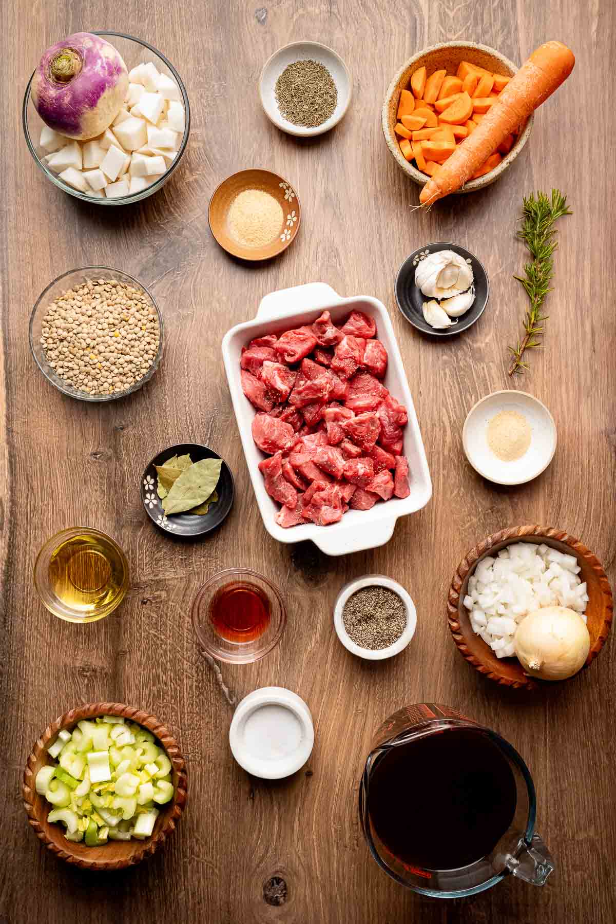 Various stew ingredients, including diced beef, chopped vegetables, seasonings, and liquids, are arranged in separate bowls and containers on a wooden surface.