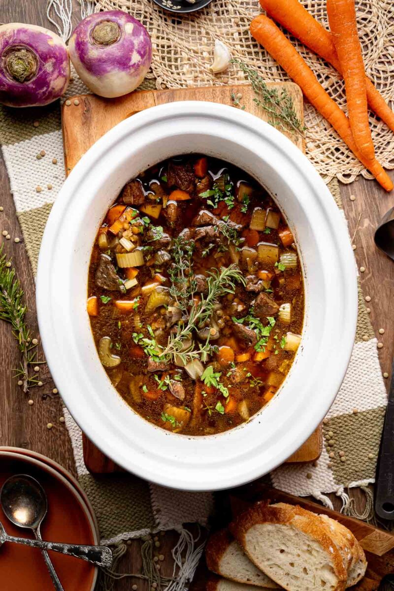 A white slow cooker filled with beef stew containing vegetables and herbs, surrounded by carrots, turnips, bread, and cooking utensils on a wooden table.