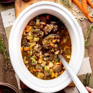 A hand stirs a vegetable and beef stew in a white slow cooker, surrounded by carrots, turnips, herbs, bread, and utensils on a wooden table.
