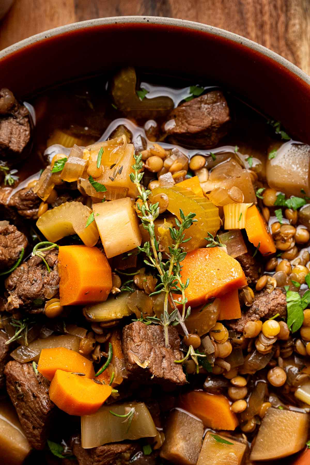 Close-up of a bowl of beef stew with chunks of beef, carrots, celery, potatoes, lentils, and garnished with a sprig of fresh thyme.