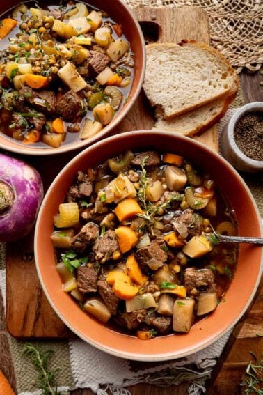 Two bowls of beef stew with vegetables and lentils are placed on a wooden board, next to slices of bread, a turnip, and a bowl of ground pepper.