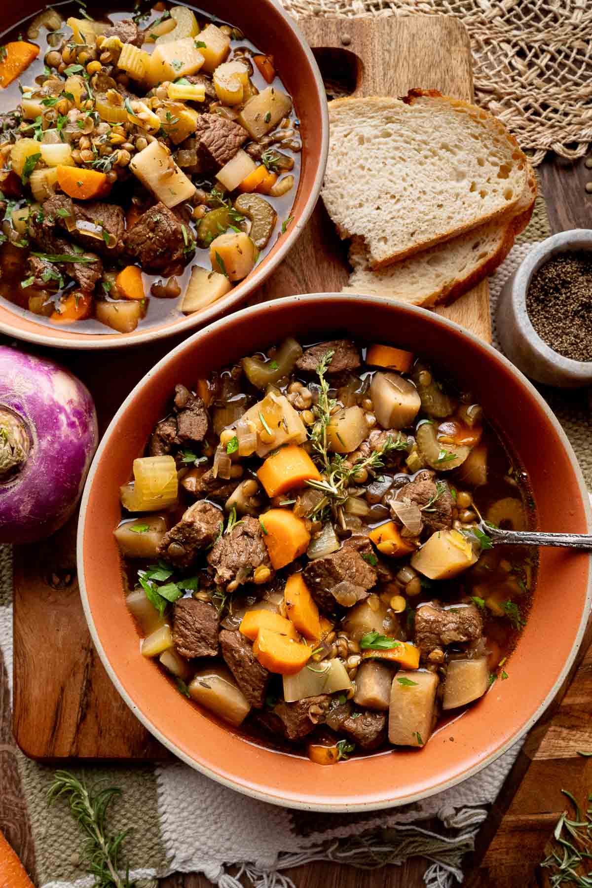 Two bowls of beef stew with vegetables and lentils are placed on a wooden board, next to slices of bread, a turnip, and a bowl of ground pepper.