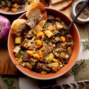 A bowl of beef stew with carrots, turnips, lentils, and bread slices, placed on a rustic wooden table with herbs and pepper nearby.