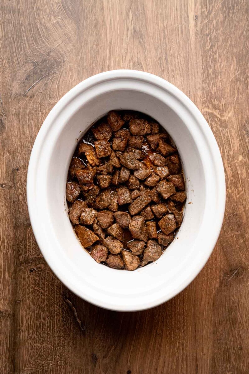 Chunks of cooked beef are placed in a white slow cooker sitting on a wooden surface.