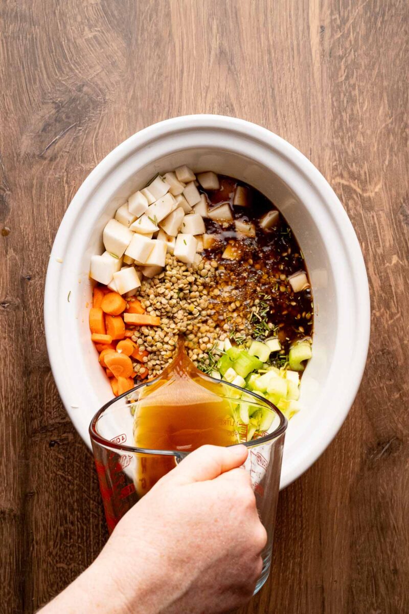 A hand pours broth from a glass measuring cup into a slow cooker filled with chopped carrots, celery, parsnips, lentils, and herbs.