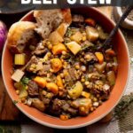 Bowl of beef stew with carrots, potatoes, and celery, topped with herbs and served with bread, placed on a rustic table setting.