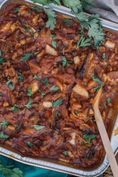 A baked casserole with a tomato-based sauce, diced potatoes, and herbs in a foil tray, garnished with fresh parsley and served with a wooden spoon on a rustic wooden table.